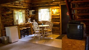 L’intérieur d’un chalet rustique comprend un coin cuisine, une table à manger, des chaises, un réfrigérateur, un téléviseur et une cheminée.