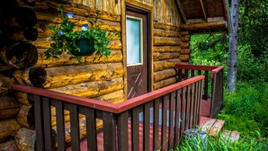 Une cabane en rondins avec un porche en bois, une porte dotée d’une fenêtre et un pot de fleurs suspendu.