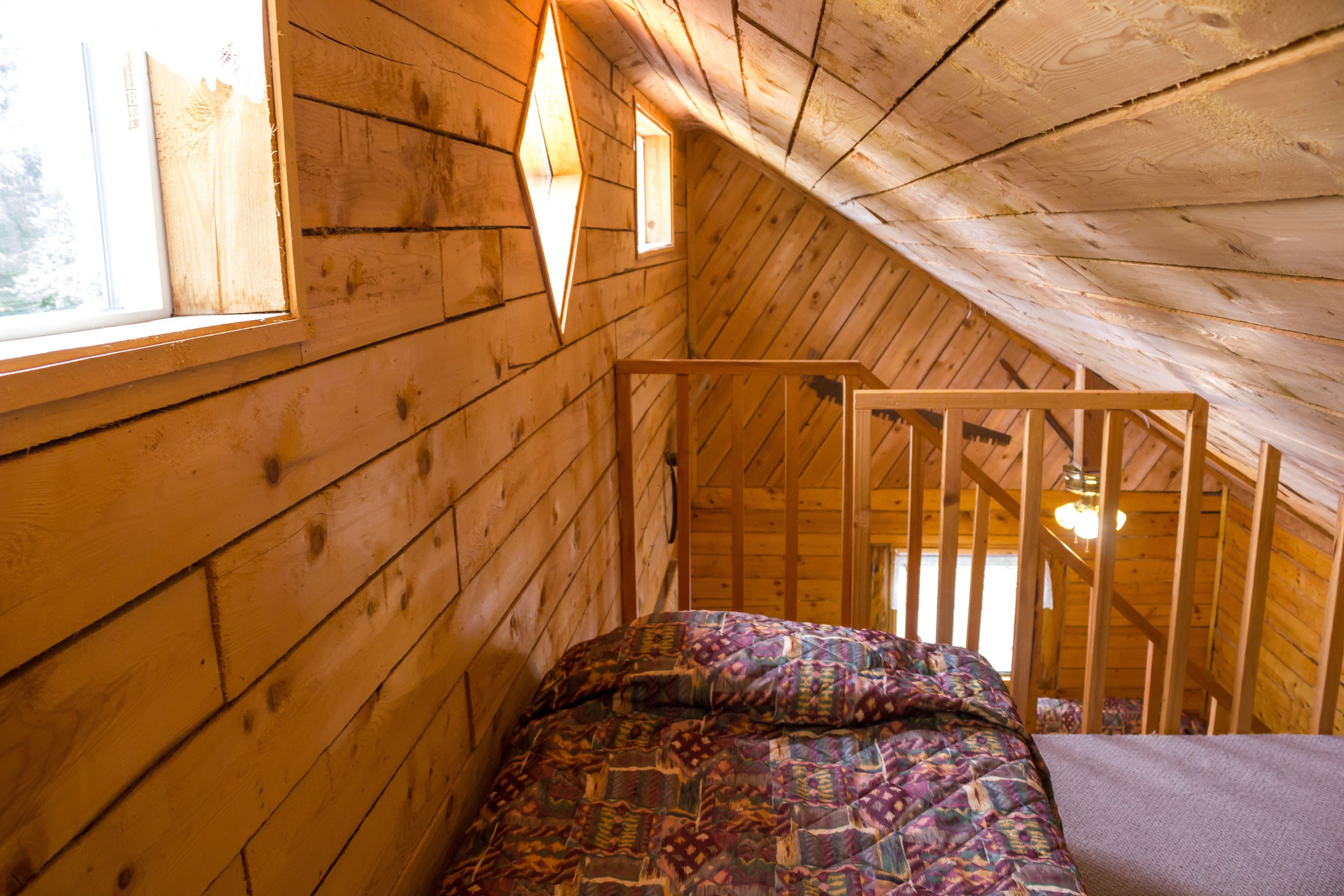 Habitación de cabaña de madera con techo inclinado, una cama con manta estampada y un ventanal con lámpara de forma de diamante.