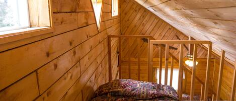 Habitación de cabaña de madera con techo inclinado, una cama con manta estampada y un ventanal con lámpara de forma de diamante.