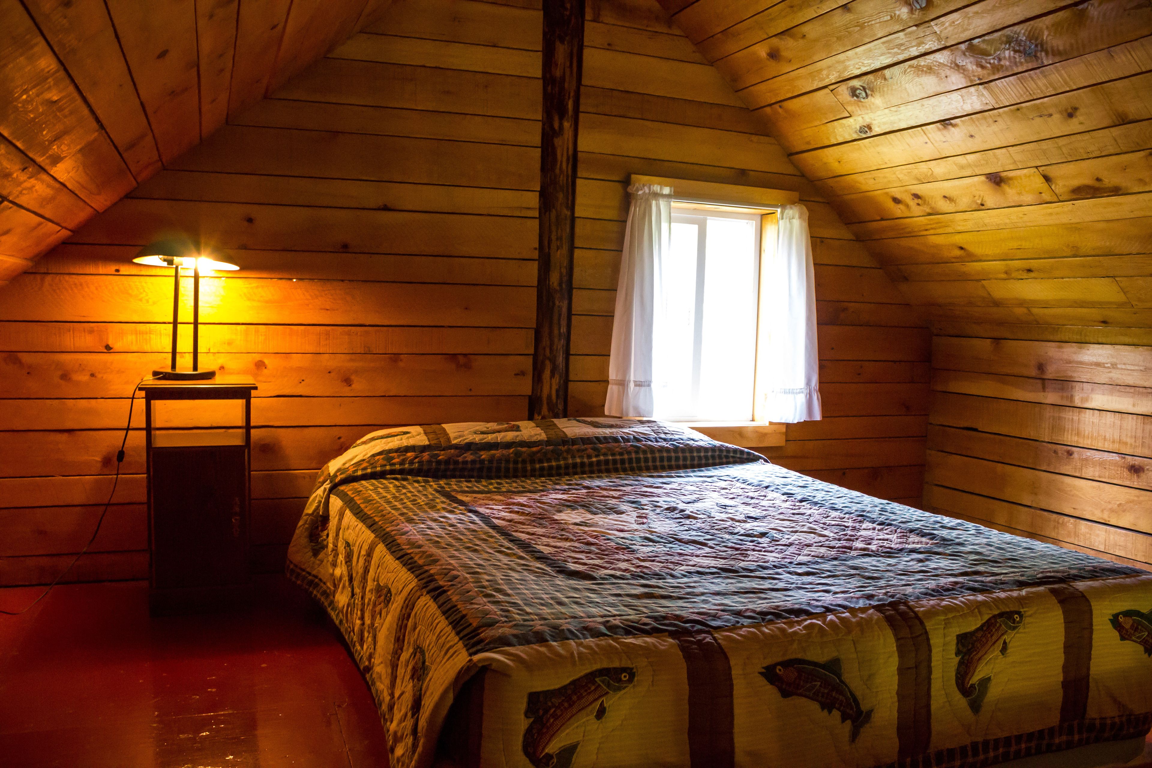 A cozy cabin room with a wooden ceiling, a bed with a quilt, a nightstand with a lamp, and a window with curtains.