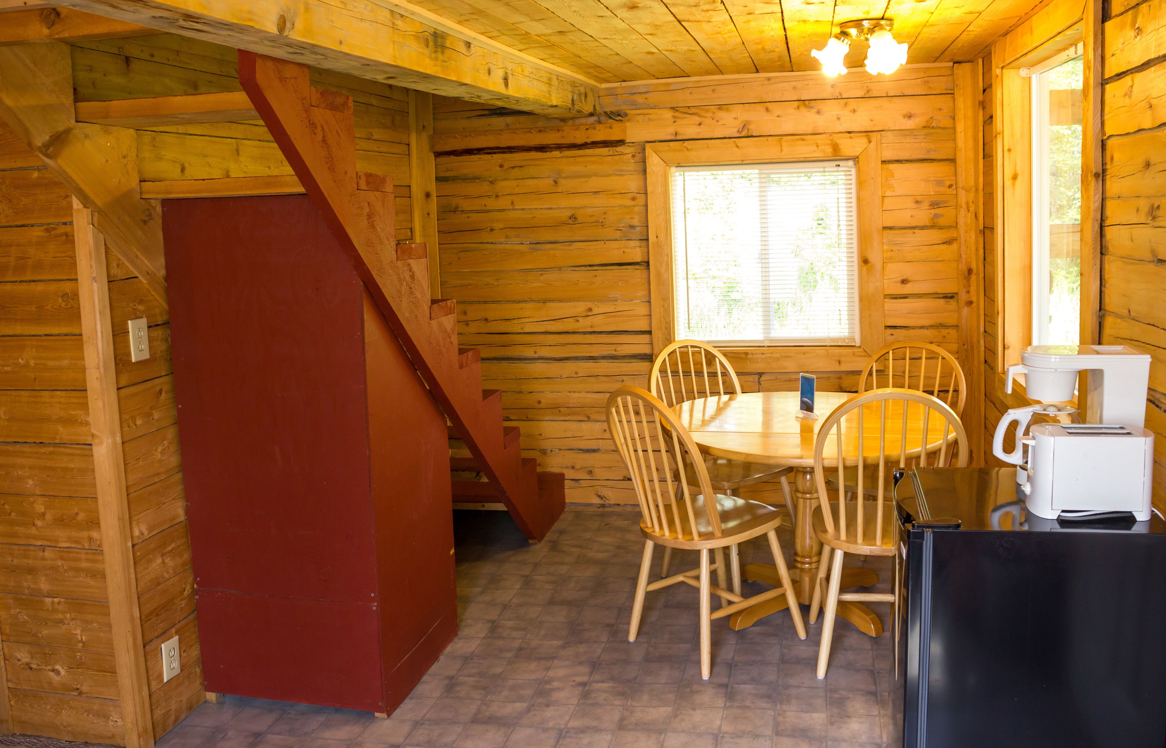 A wooden cabin interior with a dining table, four chairs, a coffee maker, a refrigerator, and a staircase.