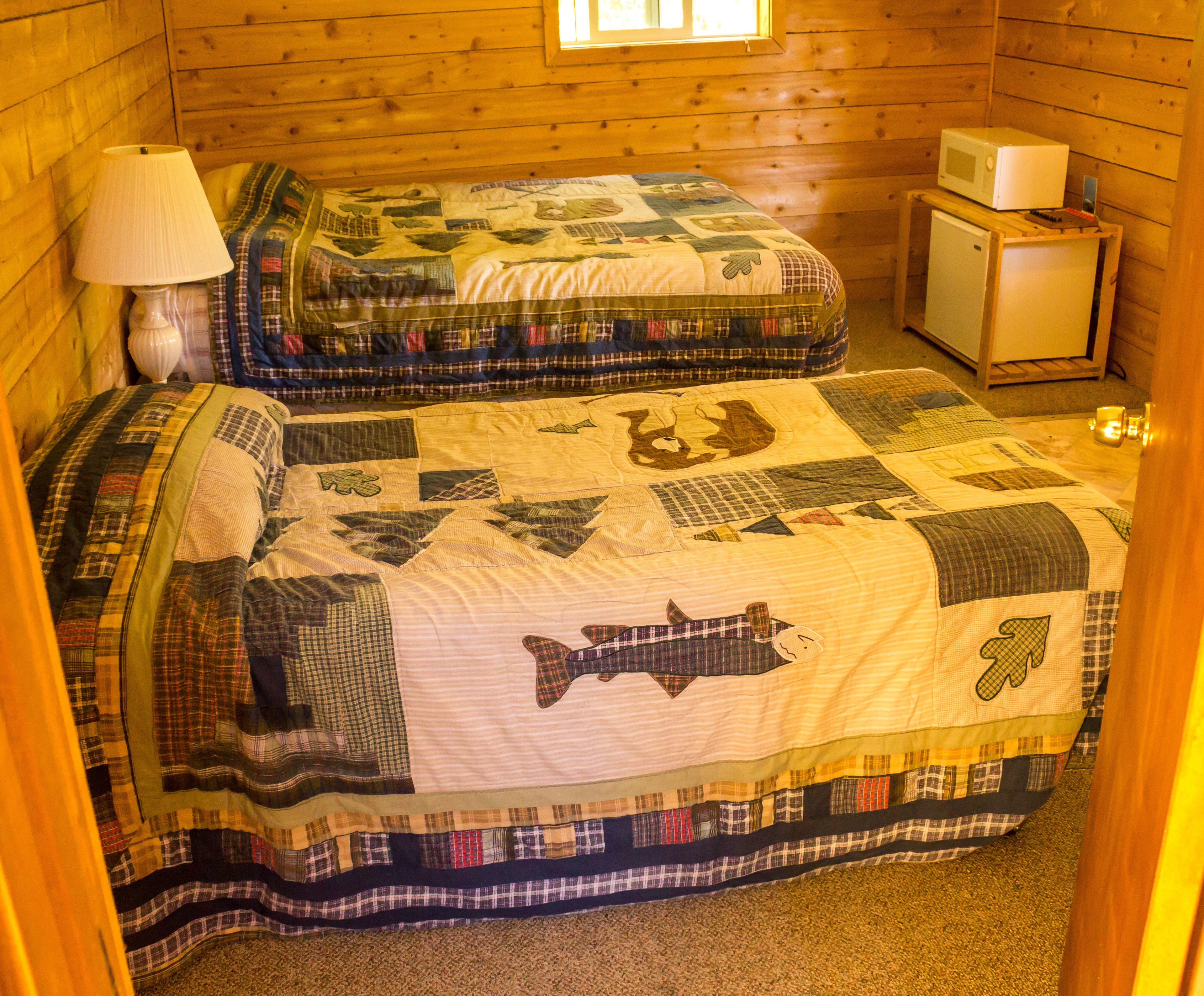 A cabin-style room with two beds, each with a quilted headboard, a small wooden nightstand with a lamp, and a microwave on a wooden shelf.