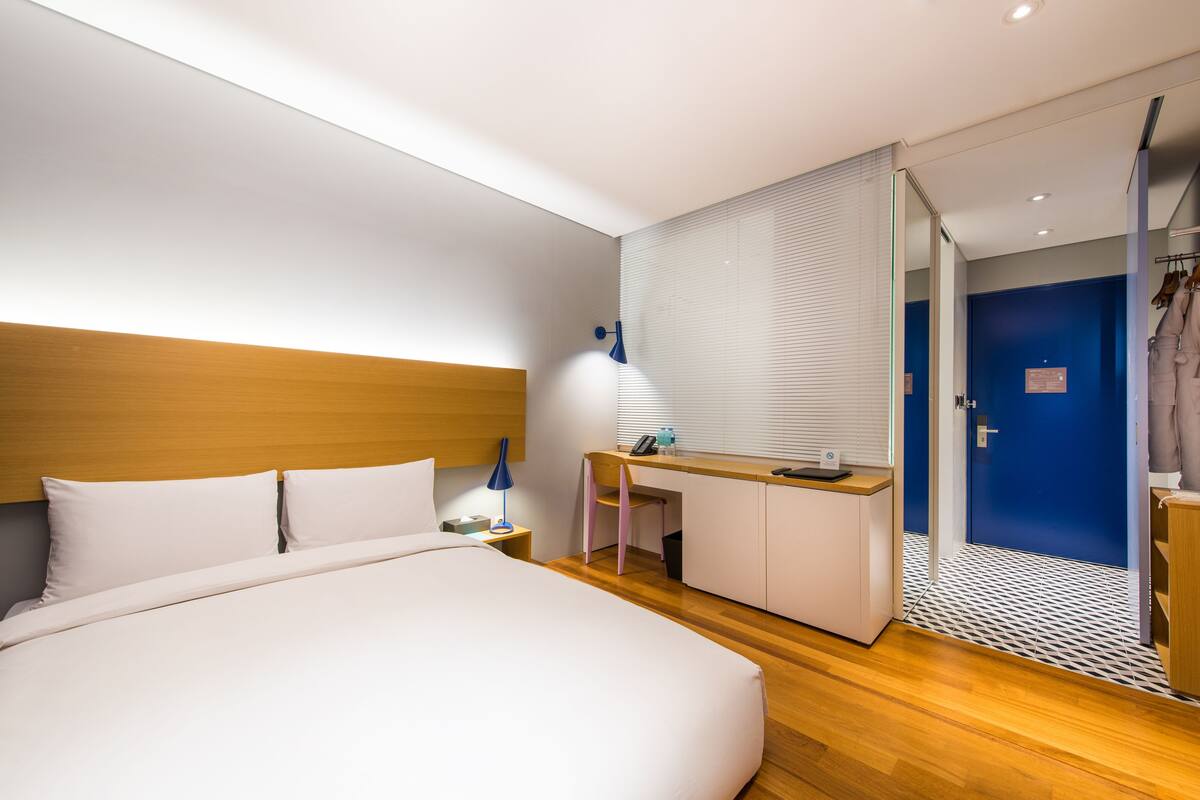 Une chambre d’hôtel moderne avec une tête de lit en bois, un grand lit, un bureau avec une chaise et un dressing avec une porte bleue.