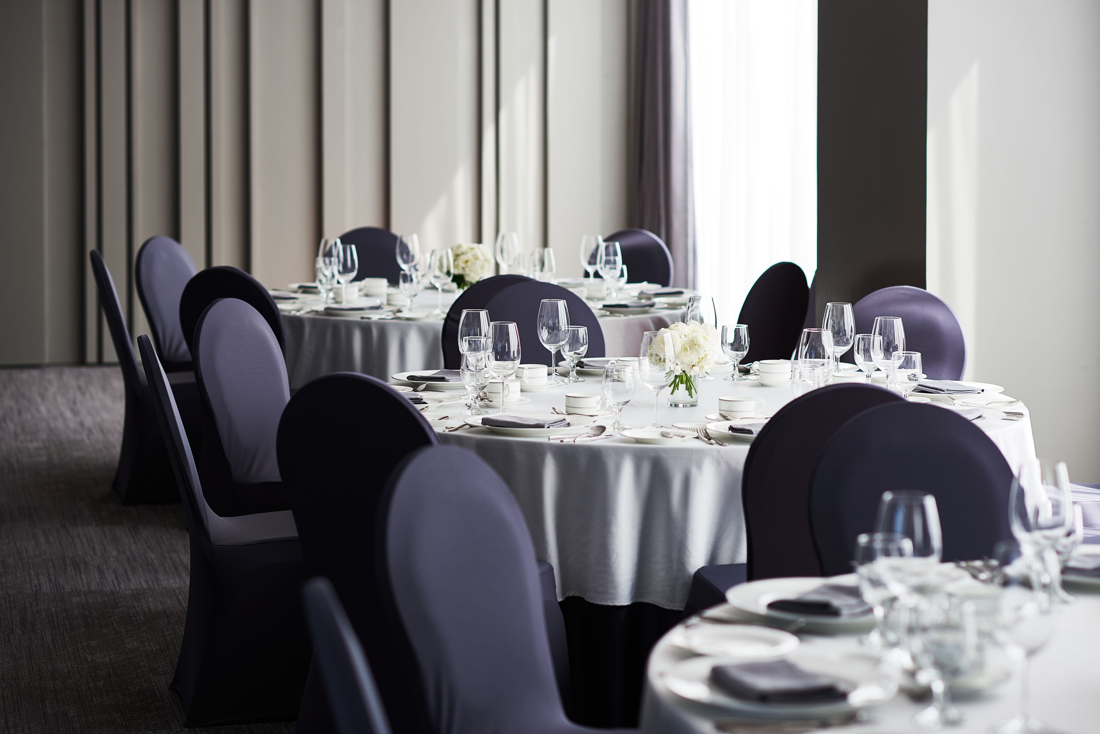 A dining area with round tables set for a formal meal, featuring white tablecloths, dark chairs, and neatly arranged glassware and plates.