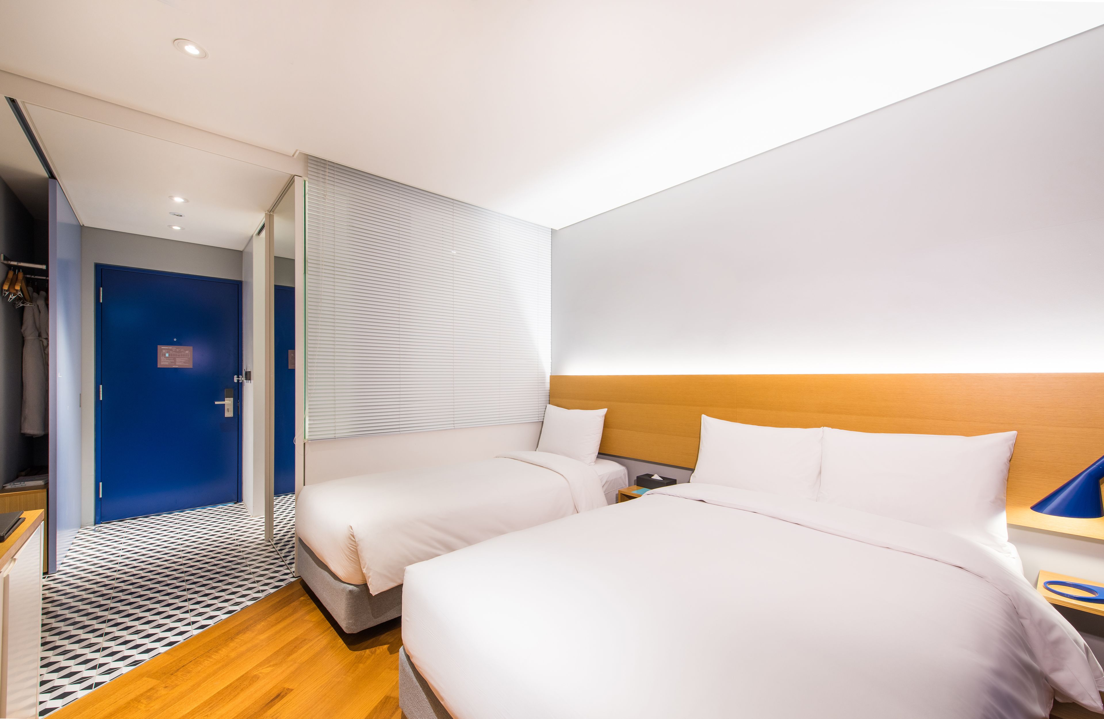A modern hotel room with two beds, a wooden headboard, a small bedside table, and a blue door.