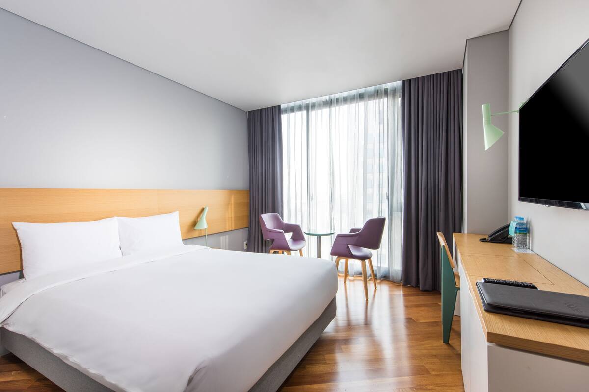 Une chambre d’hôtel avec un grand lit, deux fauteuils violets, une petite table, un téléviseur et un bureau avec un ordinateur portable et une bouteille d’eau.