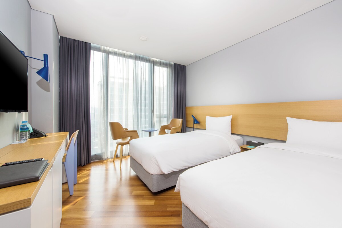 Ett hotellrum med två sängar, ett skrivbord med en bärbar dator, en tv och ett fönster med gardiner.