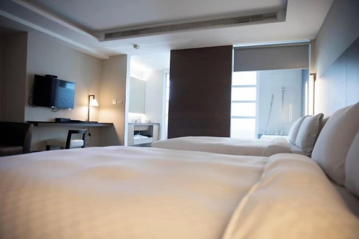 A hotel room with two beds, a desk, a flat-screen TV, and a large window.