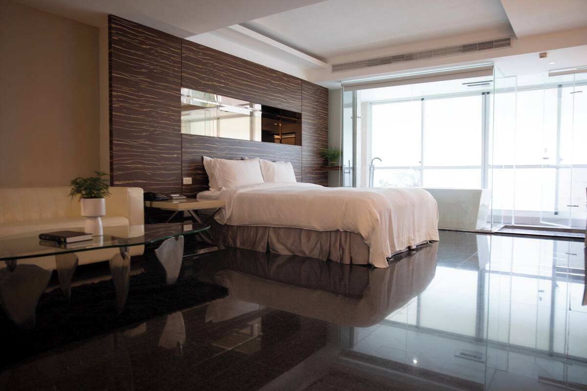 A modern bedroom with a large bed, a glass coffee table, a sofa, and a large window offering a view of the outdoors.