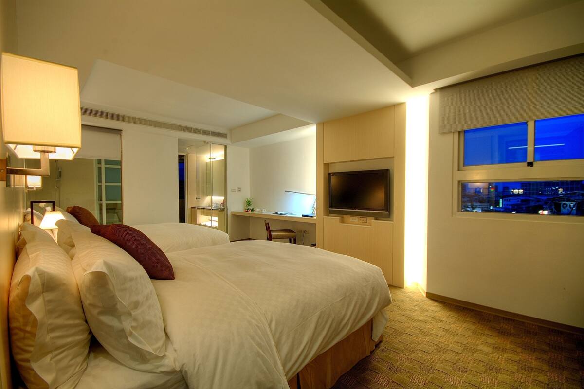 A hotel room with a large bed, a desk with a computer, a TV, and a window with a city view.