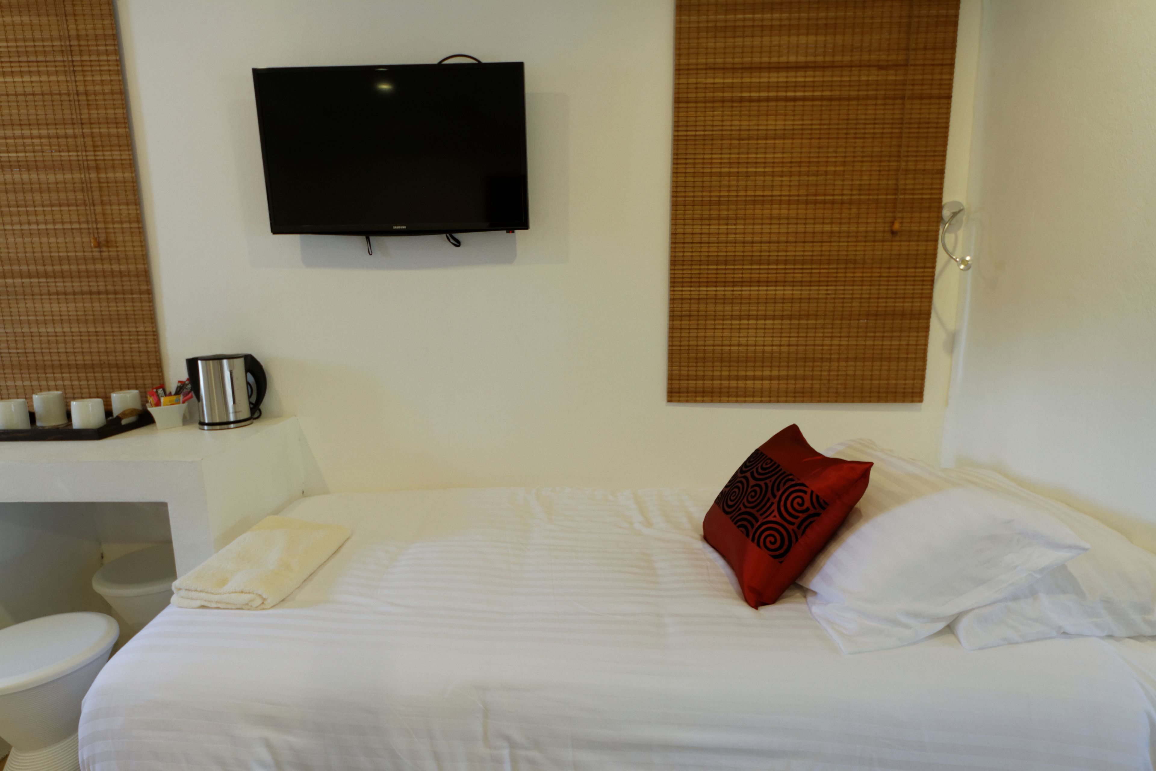 Habitación de hotel con cama, televisor montado en la pared, cafetera y ventana con persianas.