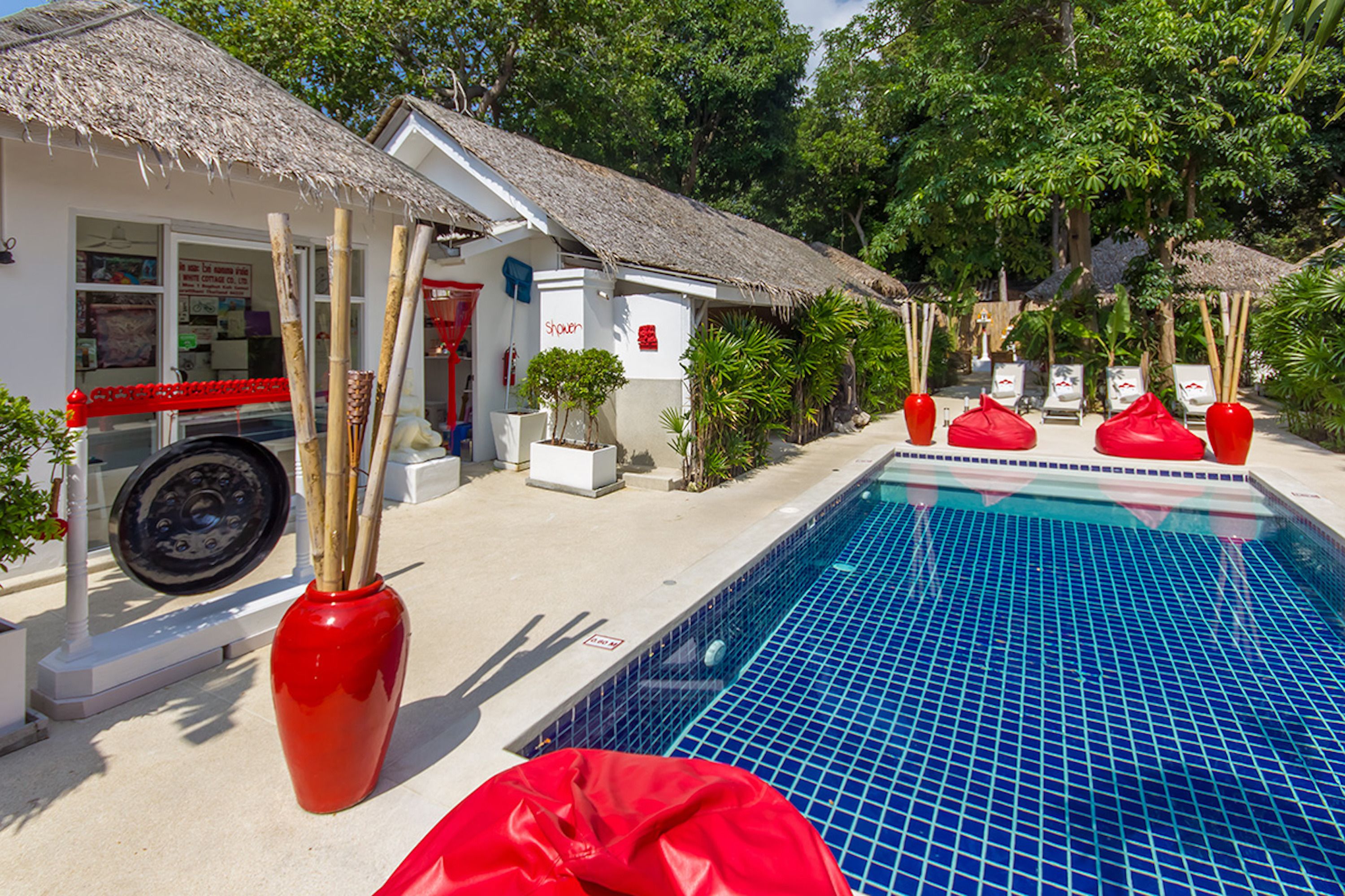 A pool area with a thatched-roof building, red vases, and lounge chairs.