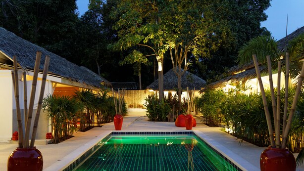 A swimming pool with a tiled floor, surrounded by plants and bamboo sticks, with a thatched-roof building in the background. - The White Cottage (Koh Samui)