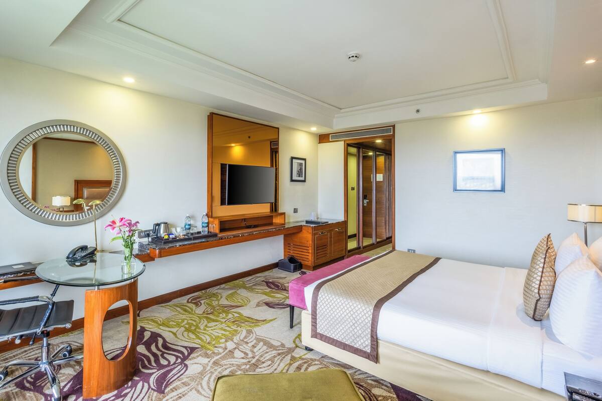 A hotel room with a bed, a desk with a chair, a TV, a mirror, and a bathroom door.