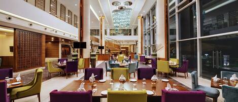A spacious hotel lobby with multiple dining areas, a grand chandelier, and a staircase leading to an upper level.