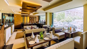 Un restaurante moderno con mesas altas, cabinas, una zona de bar y ventanas amplias con vistas a zonas verdes.