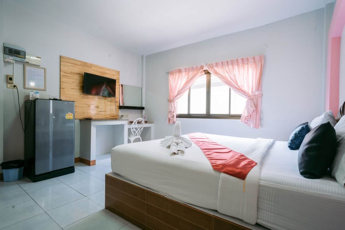A hotel room with a bed, a TV mounted on a wooden panel, a refrigerator, a desk, and a window with curtains.