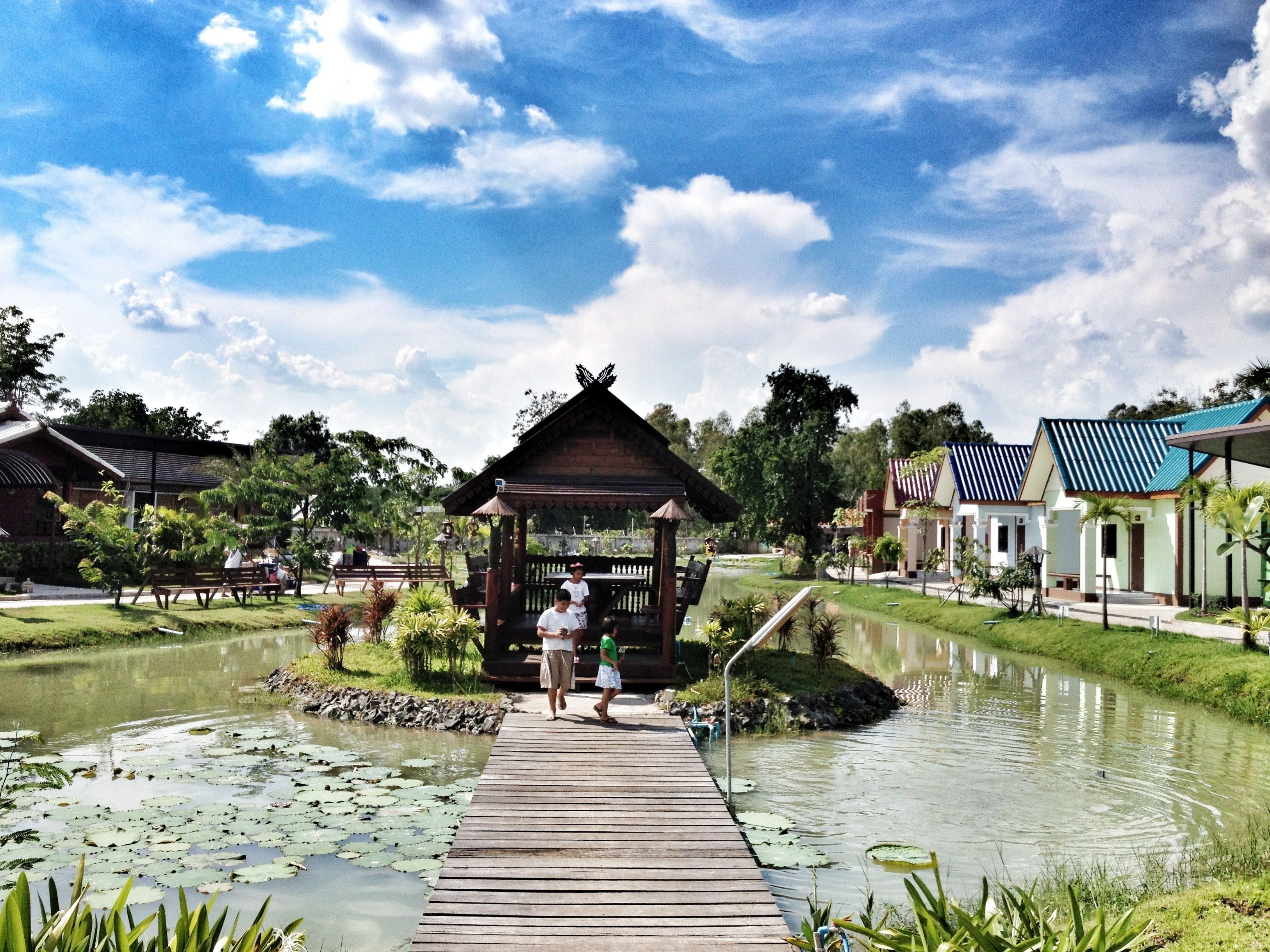 A wooden pier leading to a gazebo with a thatched roof, surrounded by a calm pond with lily pads, and buildings with blue roofs in the background.