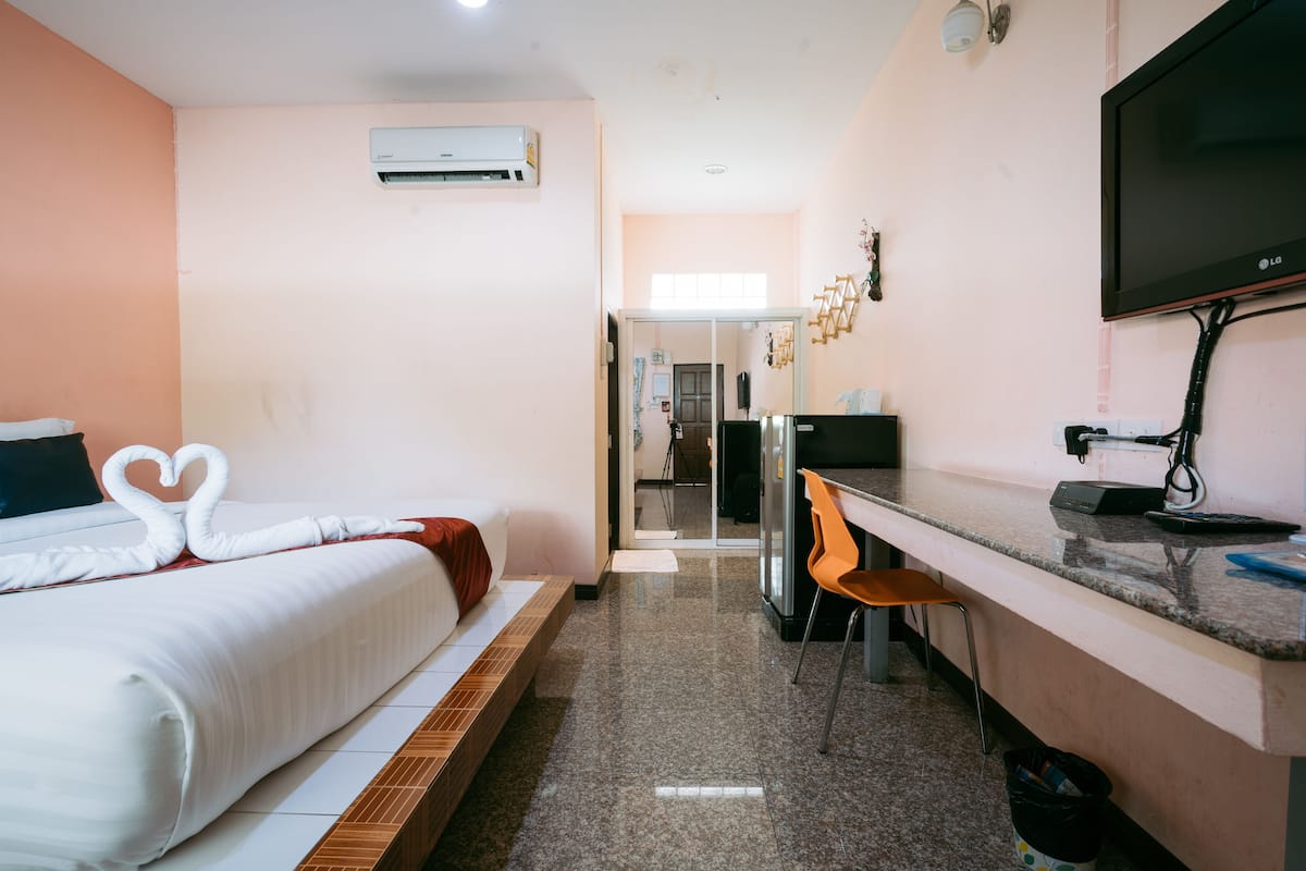 A hotel room with a bed, a desk, a chair, an air conditioner, a TV, and a trash bin.