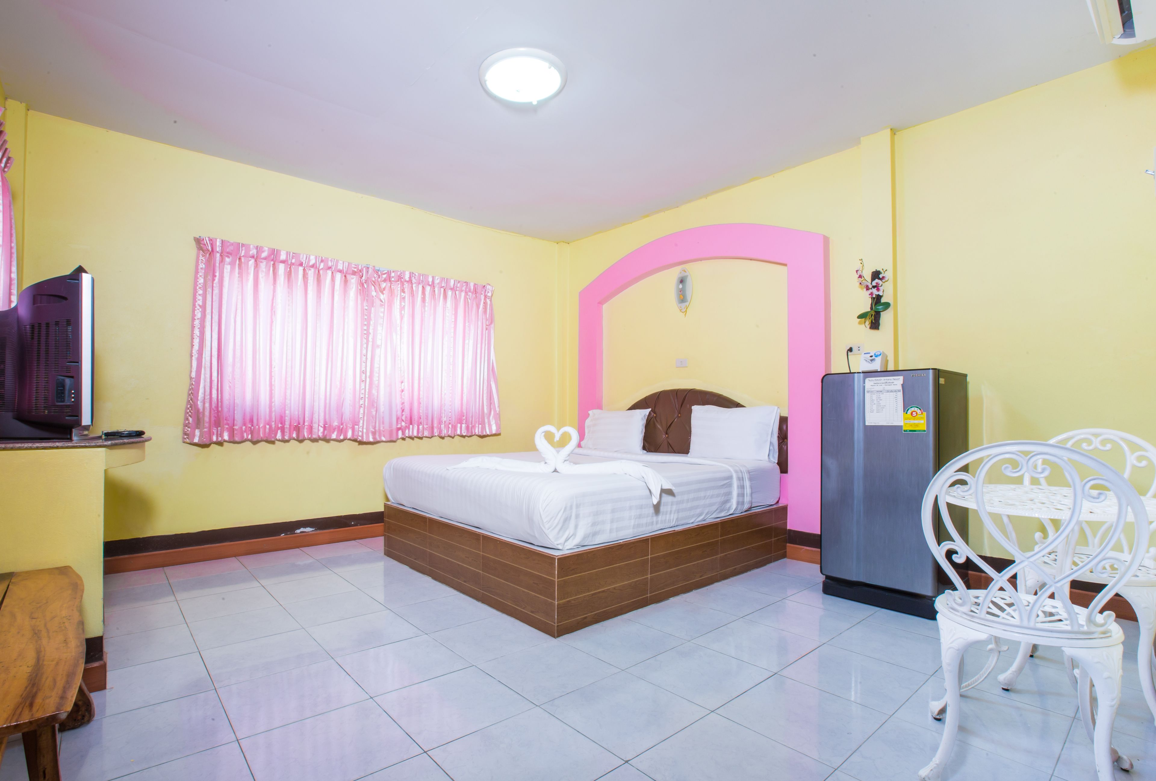 Habitación de hotel con cama, escritorio, silla, refrigerador y ventana con cortinas rosadas.