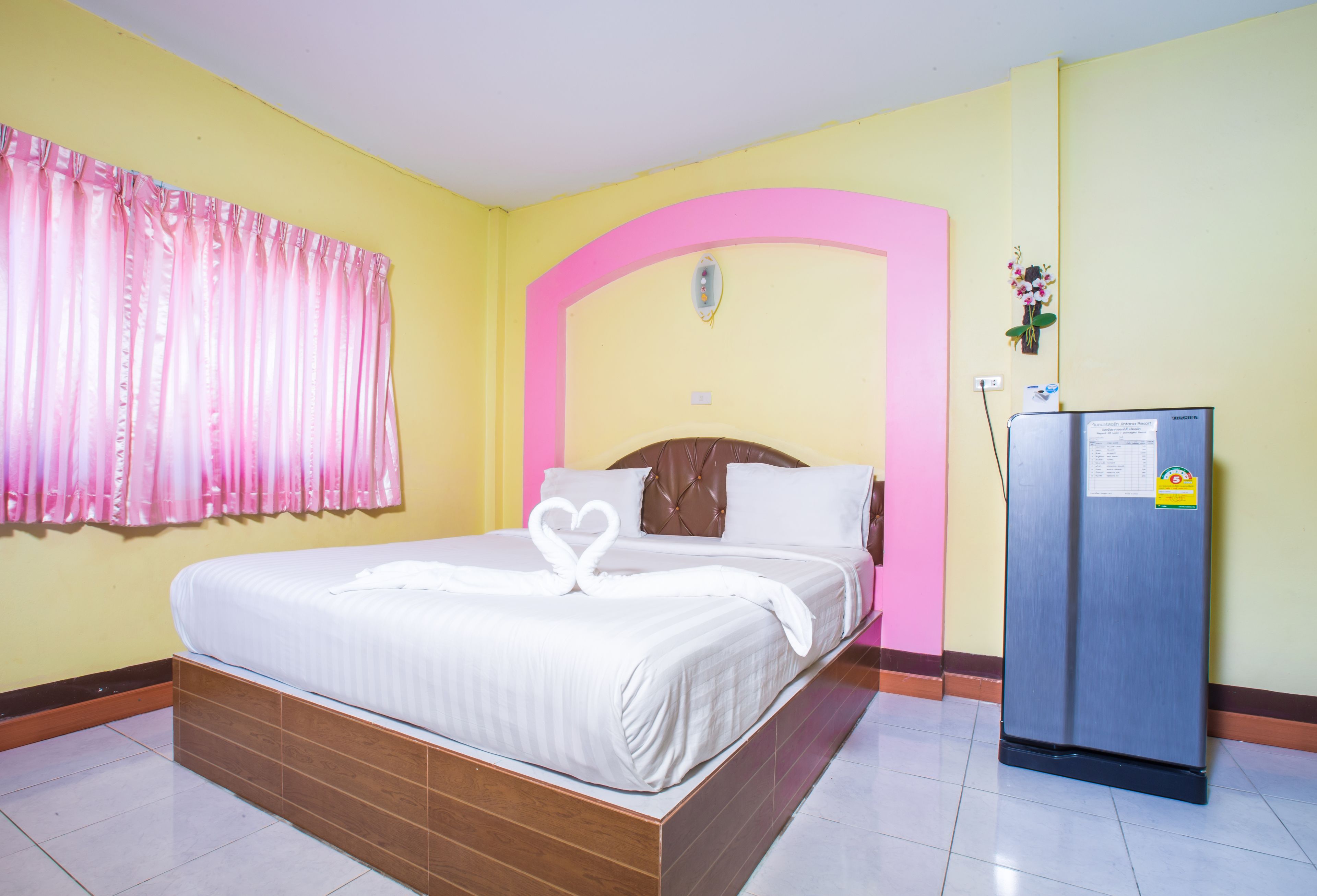 Een hotelkamer met een bed, een boog met een roze lijst, een koelkast en een raam met roze gordijnen.