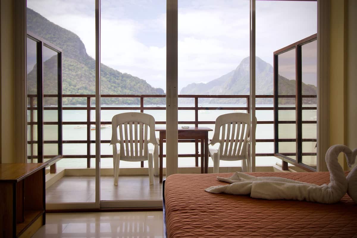 Une pièce comprenant un lit, deux chaises blanches, une table et une vue sur un plan d’eau et des montagnes, visible à travers de grandes fenêtres.