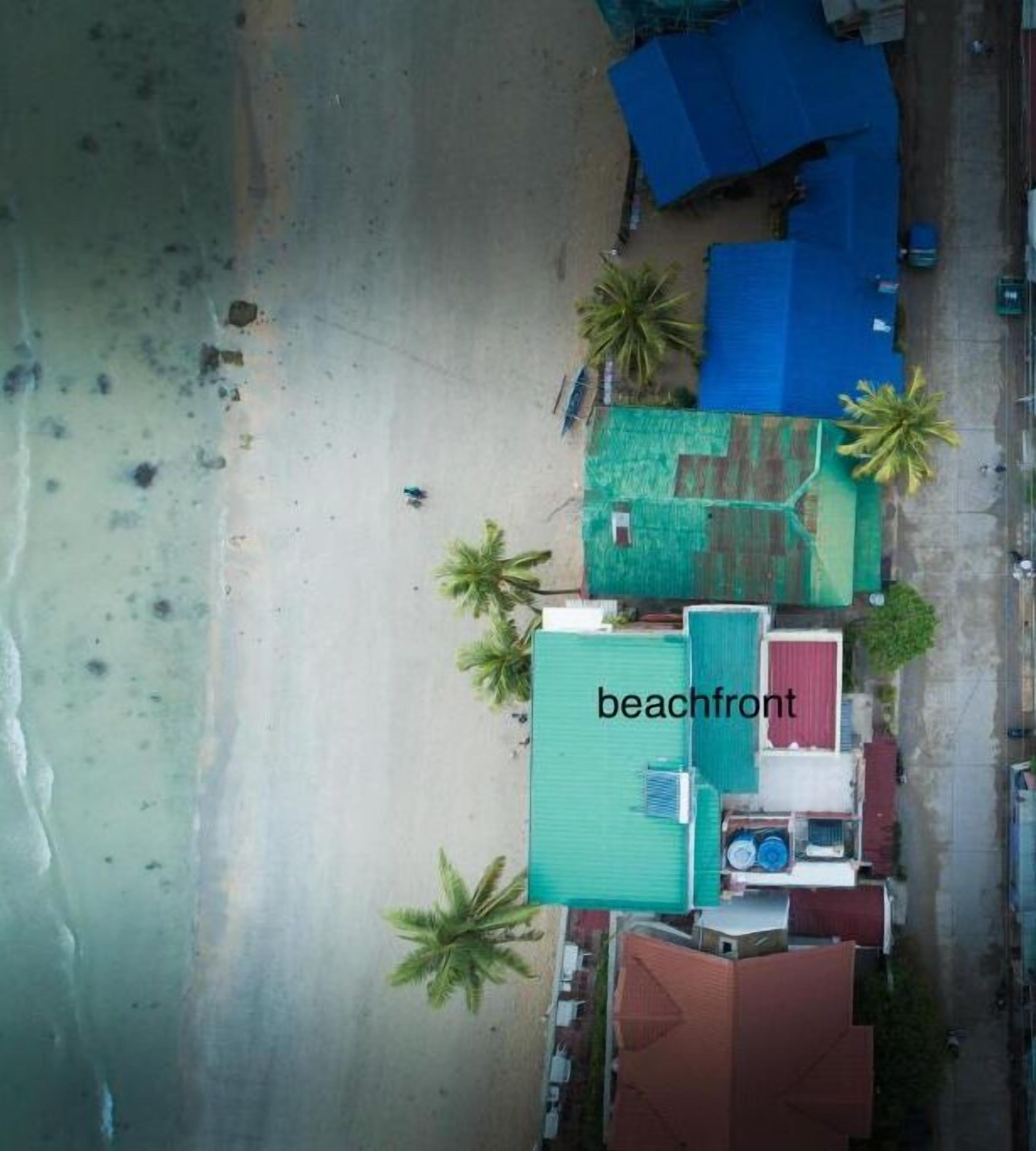 Vista aérea de uma propriedade à beira-mar com um edifício de telhado verde identificado como 'beachfront', palmeiras e uma praia de areia.