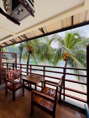 A wooden deck with a table and chairs, overlooking a beach with palm trees and boats.