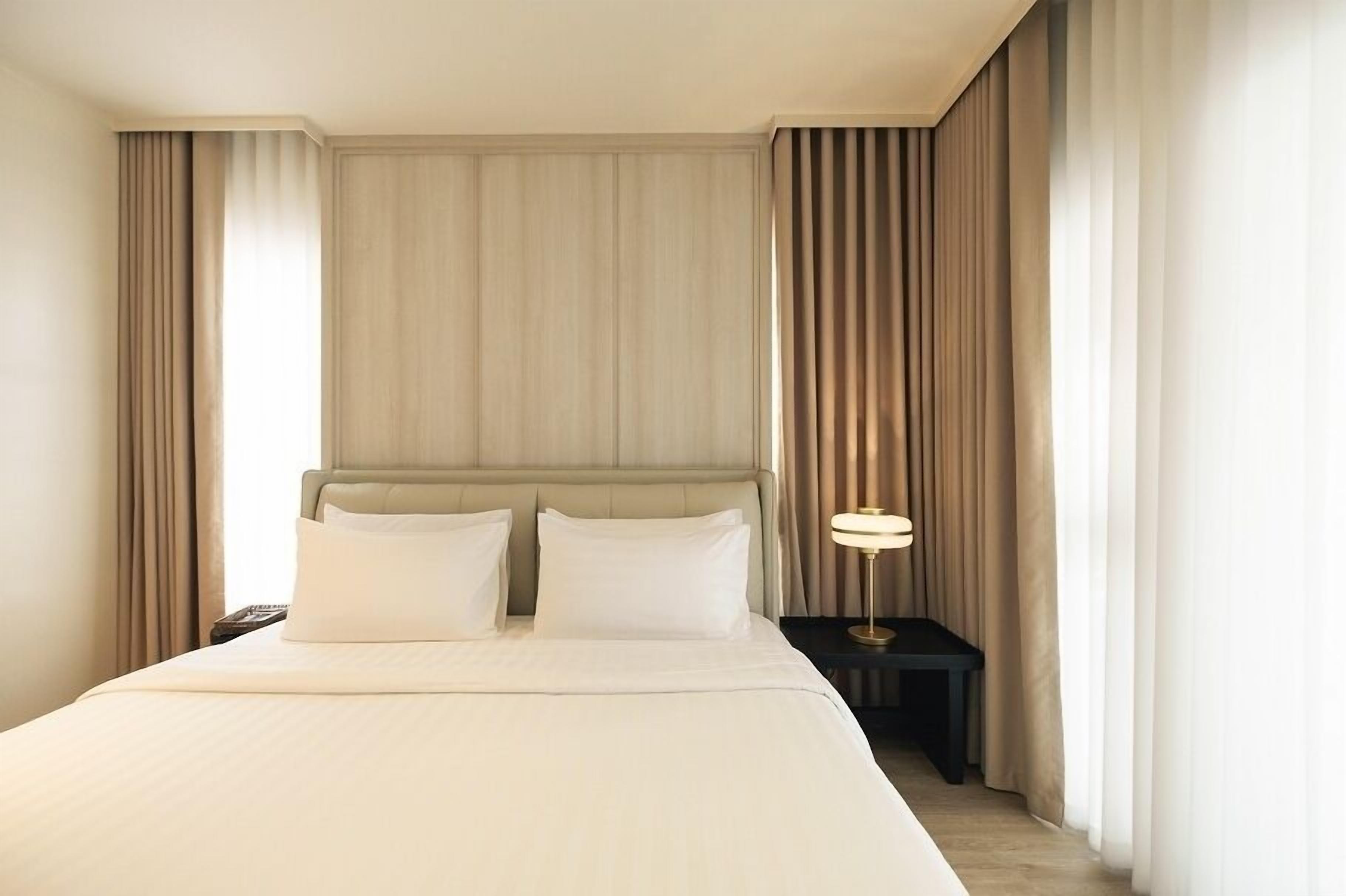 A hotel room with a large bed, two pillows, a nightstand with a lamp, and curtains on the windows.