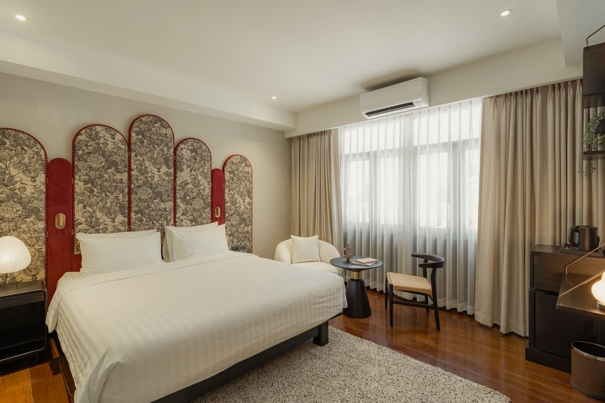 A modern hotel room with a large bed, a small round table, a chair, and a desk with a lamp.