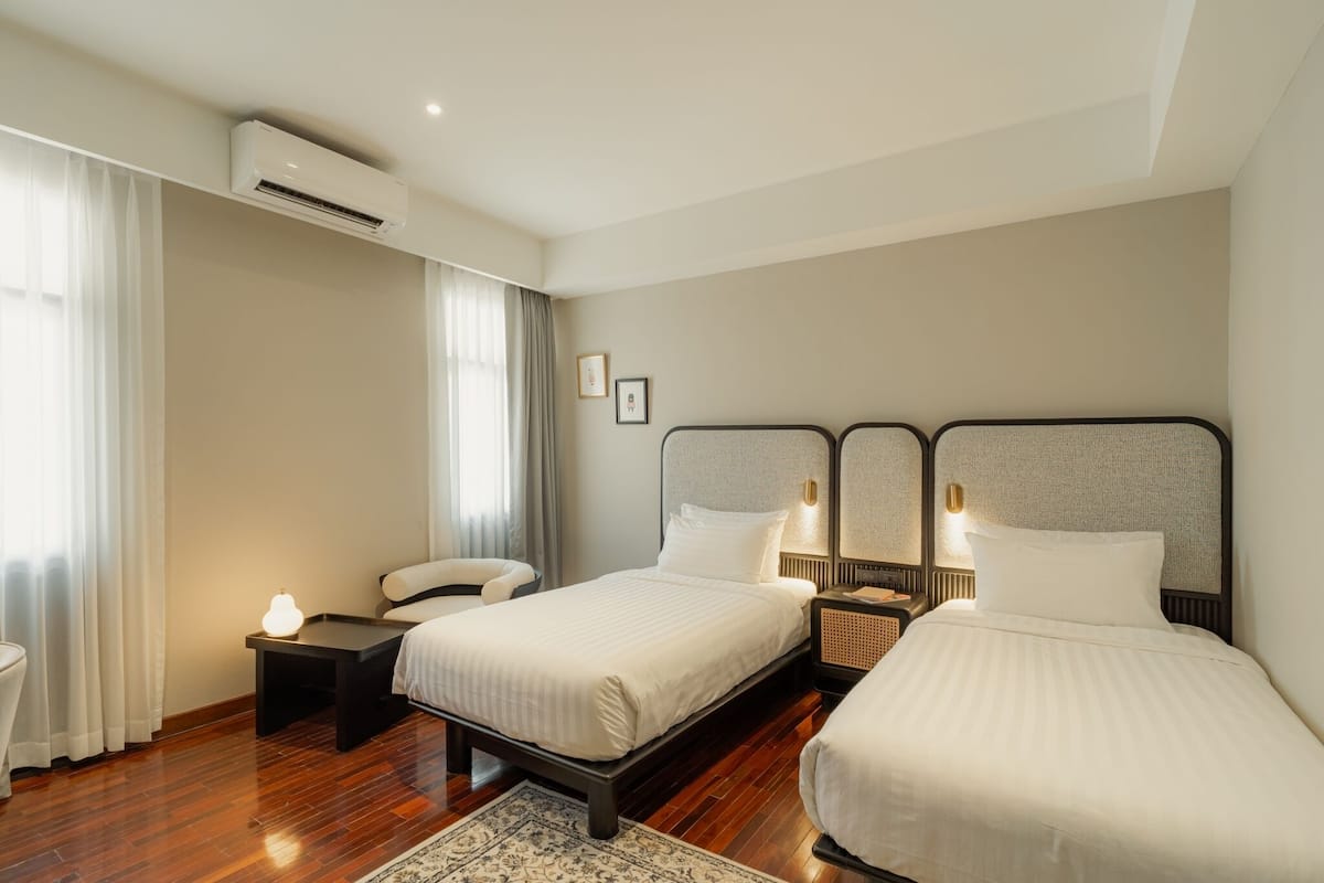 A hotel room with two beds, a bedside table, a nightstand, an air conditioning unit, a window with curtains, and a wall-mounted light fixture.