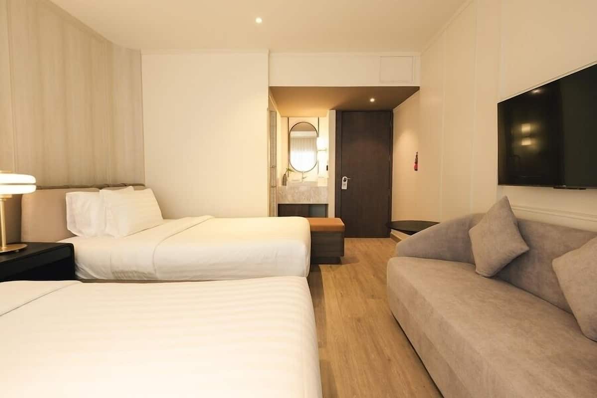 A hotel room with two beds, a sofa, a TV, a mirror, and a bathroom with a sink and toilet.