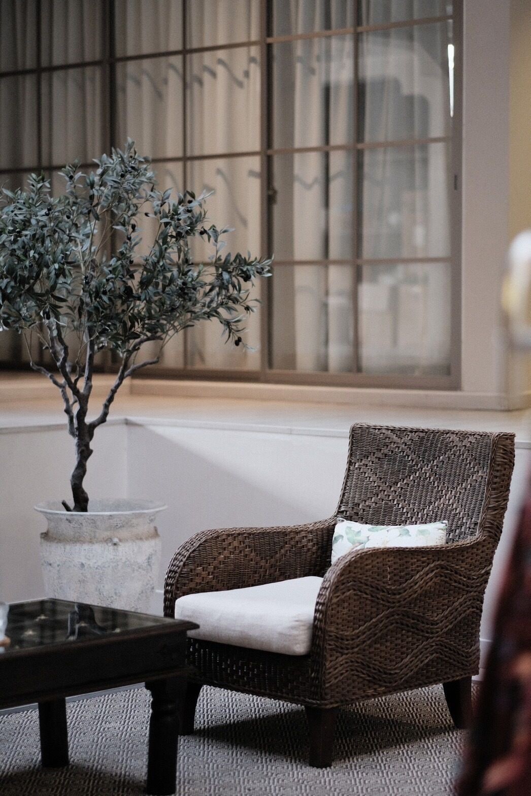 A wicker chair with a cushion, a bonsai tree, and a glass table in a room with a large window and a textured wall.