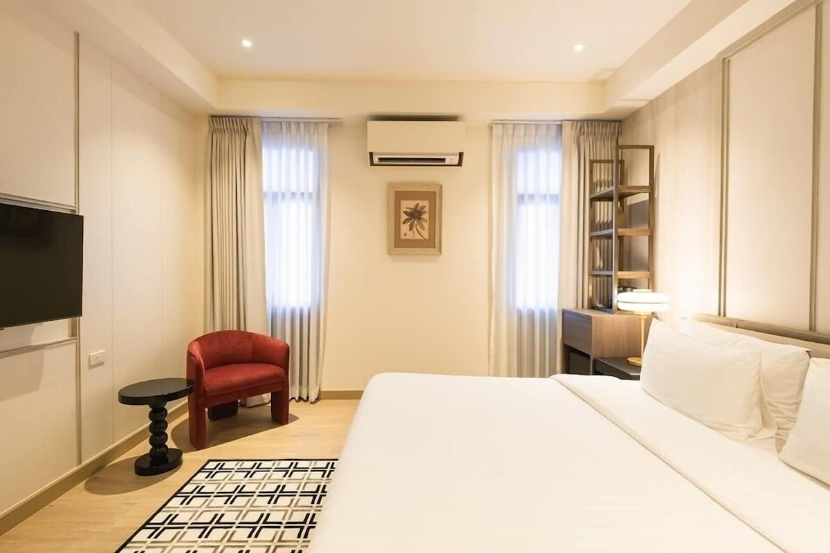 A hotel room with a bed, a red chair, a small table, a TV, a bookshelf, and a picture on the wall.