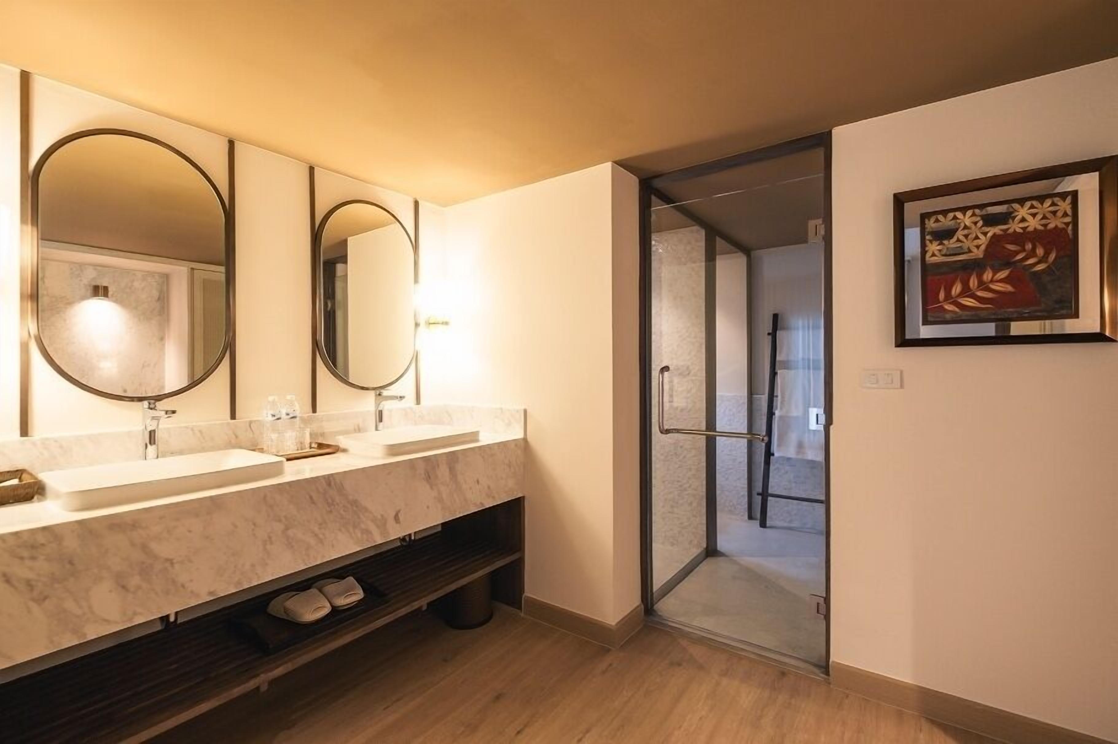 Une salle de bain moderne avec un meuble-lavabo en marbre, deux grands miroirs, une douche en verre et une œuvre d’art encadrée au mur.