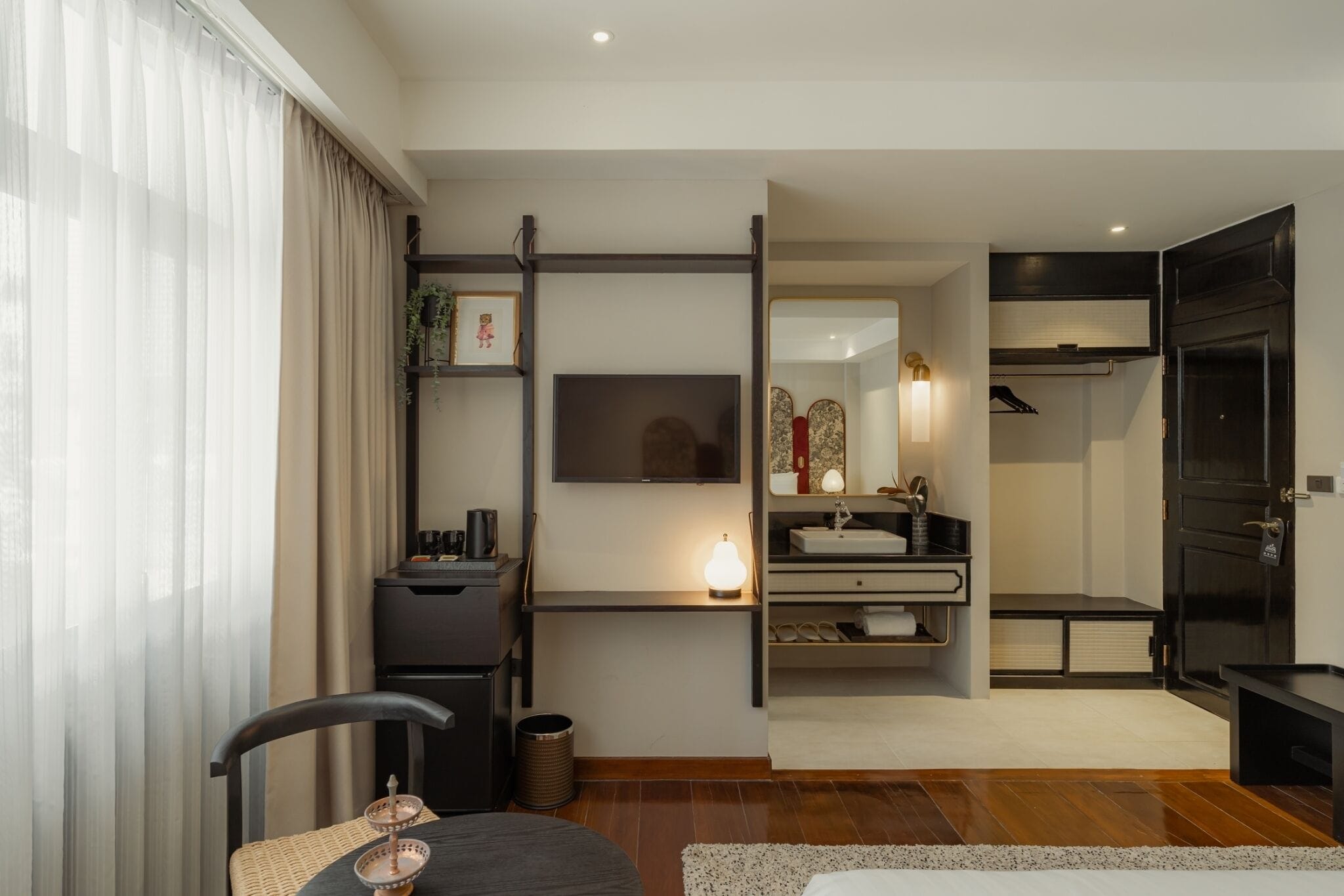 Una habitación de hotel moderna con un ventanal grande, un televisor de pantalla plana, una mesita de noche, una silla y una pequeña mesa con lámpara.