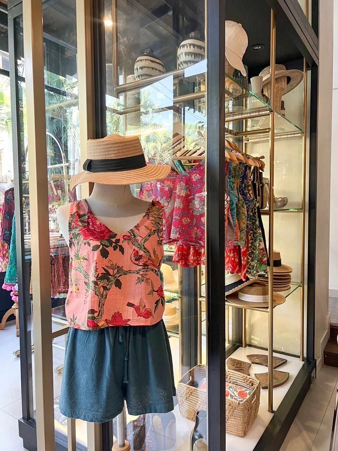 Un maniquí en un escaparate vestido con una blusa y pantalones cortos estampados con flores, con sombreros y accesorios exhibidos al fondo.