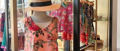 Un maniquí en un escaparate vestido con una blusa y pantalones cortos estampados con flores, con sombreros y accesorios exhibidos al fondo.