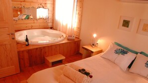 A wooden cabin bedroom with a large bed, a wooden bathtub, a bedside table with a lamp, and a mirror on the wall.