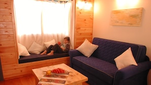 A cozy cabin interior with a wooden floor, a sofa, a small table with a fruit bowl, and a window with curtains.