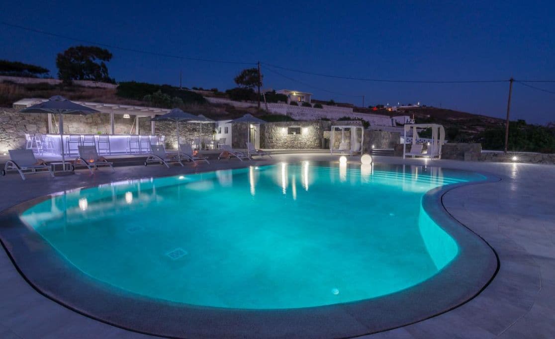 Una piscina con sillas de descanso y sombrillas, rodeada de muros de piedra y un edificio de piedra con luces.