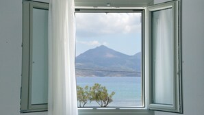 Una habitación con ventana abierta que muestra una montaña y un árbol junto al agua.