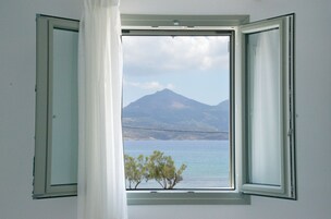 Una habitación con ventana abierta que muestra una montaña y un árbol junto al agua.