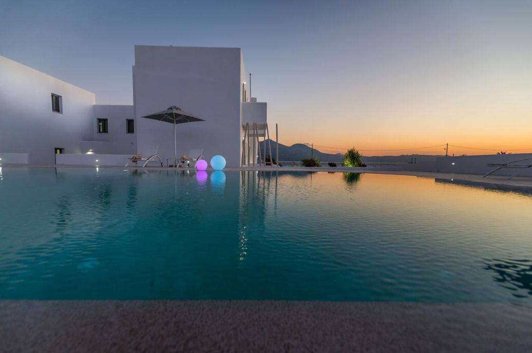 Un área de piscina moderna con vista despejada a un edificio blanco y un atardecer al fondo.