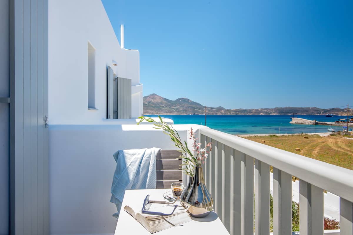 A balcony with a table set for two, a vase with flowers, and a view of the sea and mountains.
