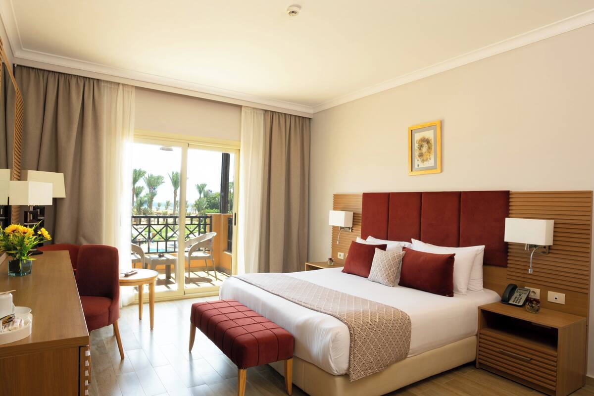 A hotel room with a bed, bedside tables, a desk, a chair, and a balcony with a view of palm trees.