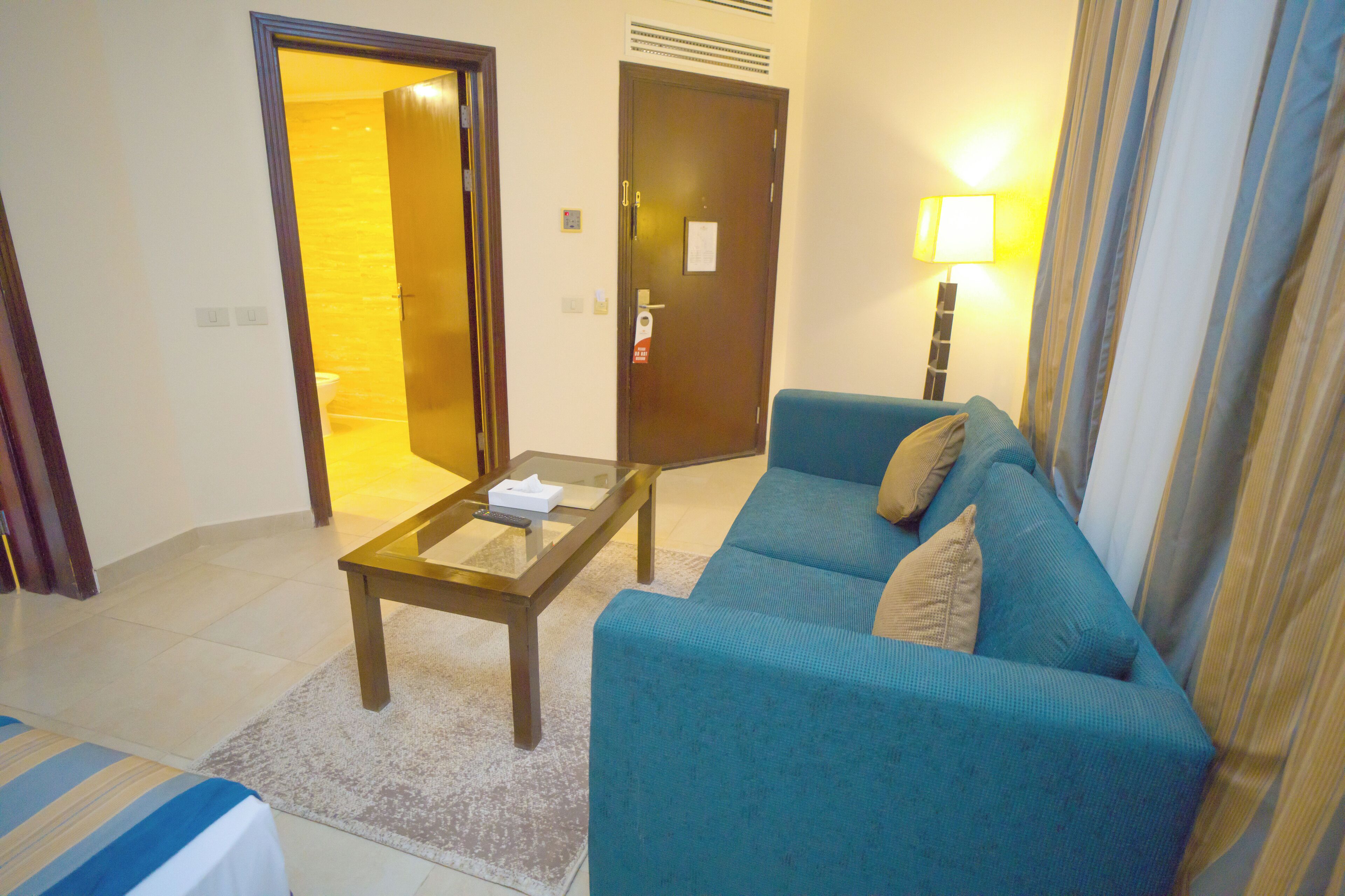 A hotel room with a blue sofa, a glass coffee table, a lamp, and a door leading to another room.