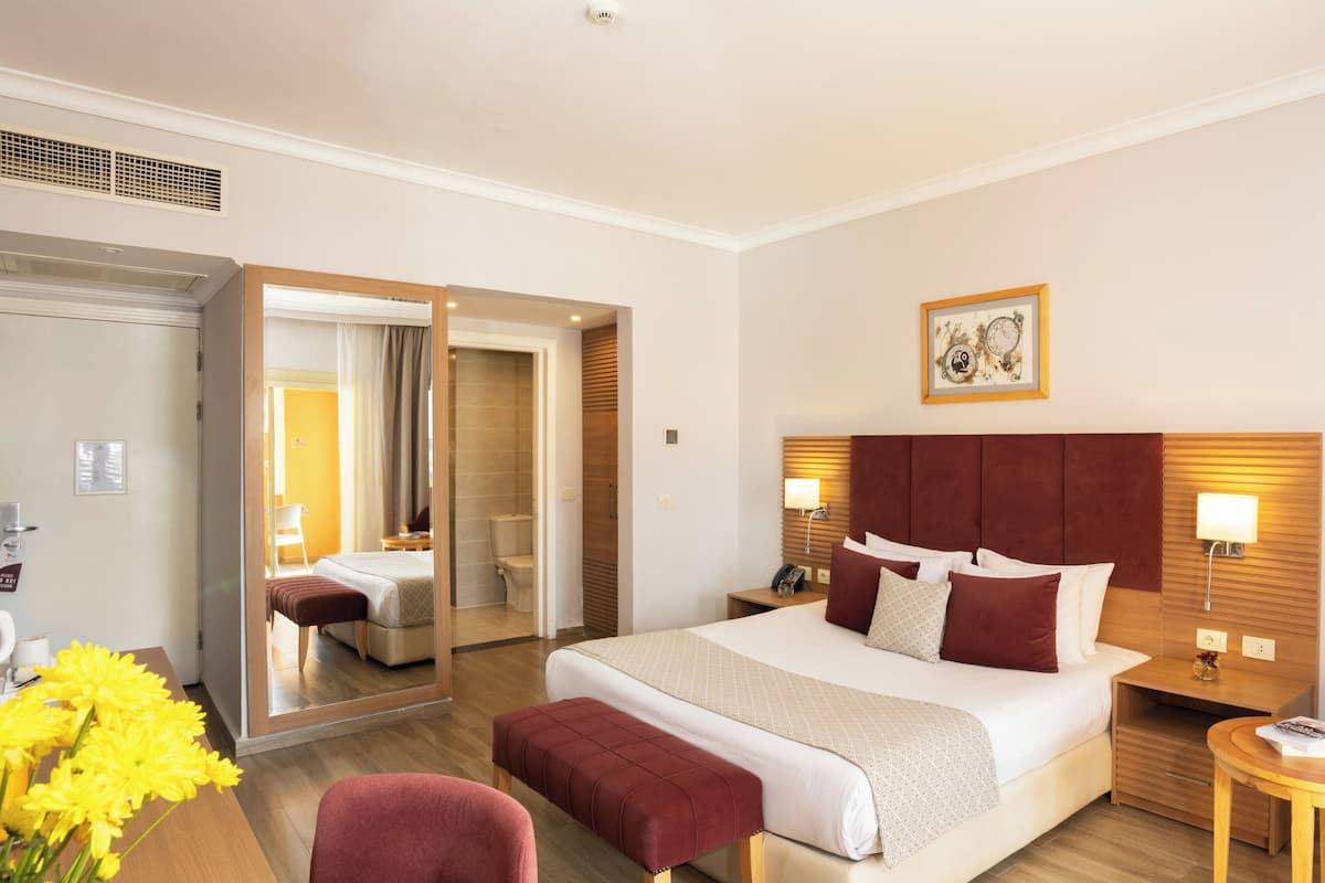 A hotel room with a large bed, a wooden headboard, a bedside table with a lamp, a mirror, and a small desk with a chair.