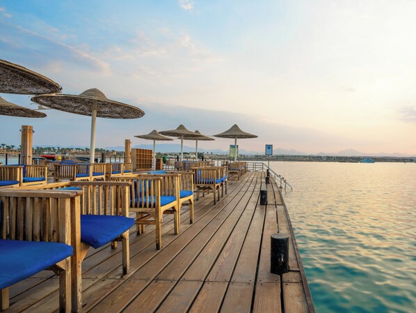 A wooden deck with blue cushioned chairs and thatched umbrellas, overlooking a calm body of water with distant mountains.